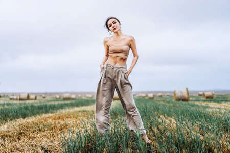 Brunette in fashionable clothes with bare shoulders standing in a wheat field on green grass. Woman holds her hands in pants pockets and looking forward.の写真素材