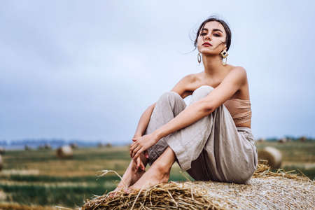 Brunette in linen pants and bare shoulders sitting on a hay bales in warm autumn day. Woman looking at camera. Behind her is a wheat field.の写真素材