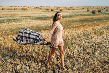 Full length portrait of a beautiful brunette in a dress and with a warm plaid. Woman enjoying a walk in a wheat field with hay bales.の写真素材