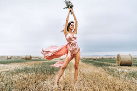 Young brunette in a pink satin dress in a wheat field near hay bales. A woman has a wreath on her head and holds a bouquet of wildflowers in her hands.の写真素材