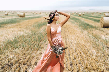 Young brunette in a pink satin dress in a wheat field near hay bales. A woman has a wreath on her head and holds a bouquet of wildflowers in her hands.の写真素材