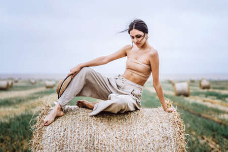 Barefoot brunette in linen pants and bare shoulders sitting on a hay bales in warm autumn day. Woman looking at camera. Behind her is a wheat field.の写真素材