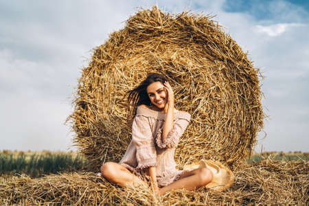 A young woman with long hair and in a dress sits near a hay bale. Woman posing smiling and looking at camera.の写真素材