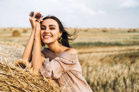 Closeup portrait of beautiful smiling woman with closed eyes. The brunette leaned on a bale of hay. A wheat field on the background.の写真素材