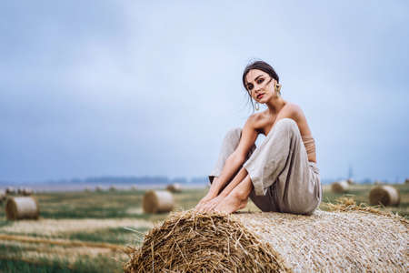 Brunette in linen pants and bare shoulders sitting on a hay bales in warm autumn day. Woman looking at camera. Behind her is a wheat field.の写真素材