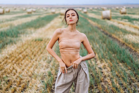 Brunette in fashionable clothes with bare shoulders standing in a wheat field on green grass. Woman holds her hands in pants pockets and looking forward.の写真素材