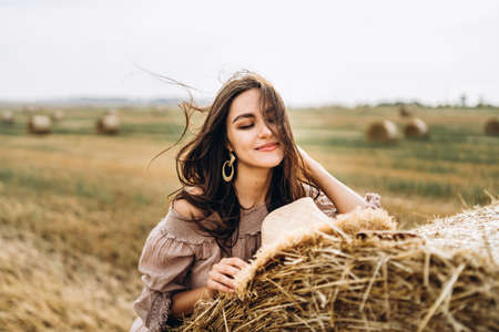 Closeup portrait of beautiful smiling woman with closed eyes. The brunette leaned on a bale of hay. A wheat field on the background.の写真素材
