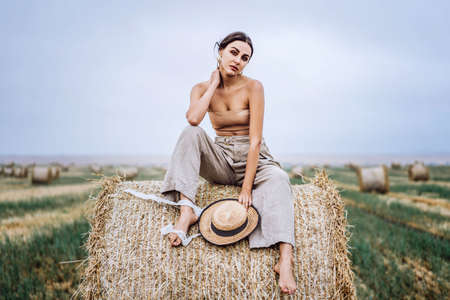 Barefoot brunette in linen pants and bare shoulders sitting on a hay bales in warm autumn day. Woman looking at camera. Behind her is a wheat field.の写真素材