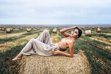 Young slender brunette girl with bright makeup in beige top with bare stomach and gray pants, lying on hay, posing for camera, photo shoot on field, blue skyの写真素材