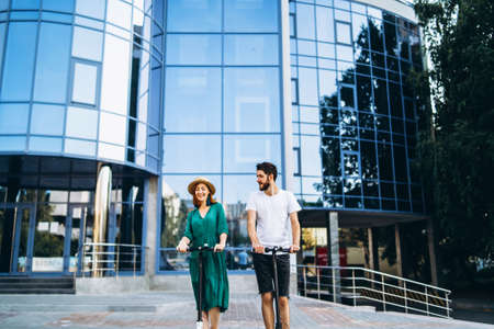 Full length portrait of a young romantic couple with electric scooters, walking in the city. Young woman in hat and man enjoy a walk.の写真素材