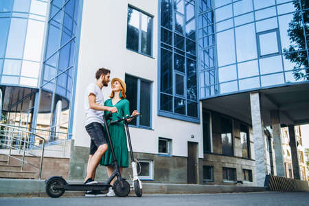 A young smiling couple enjoy a walk in the city on electric scooters. Content technologies. Large modern building on background.の写真素材