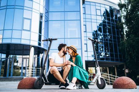 Young loving couple relaxing sitting near a modern glass building with their electric scooters. Walks in the big city.の写真素材