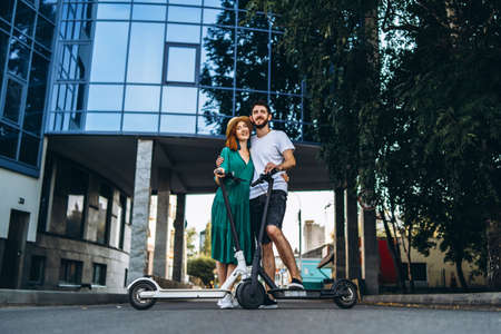 Full length portrait of a young romantic couple with electric scooters on a date, walking in the city. Young woman in hat and man enjoy a walk.の写真素材
