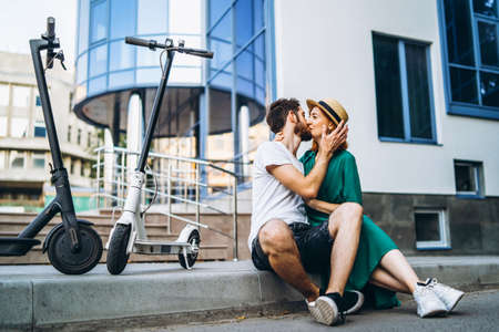 Young loving couple relaxing sitting near a modern glass building with their electric scooters. Walks in the big city.の写真素材