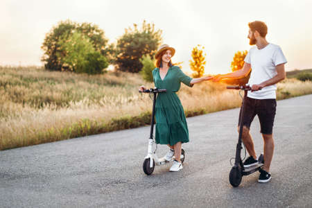 Man and woman holding hands. On the background road and sunset. Walking on electric scooters.の写真素材