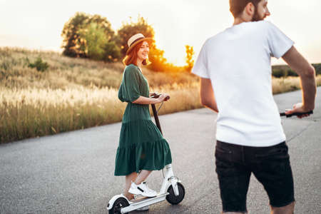 Happy couple walking on scooters. A young woman in dress and hat on sunset background. In the foreground, a man turns his back on the lens.の写真素材