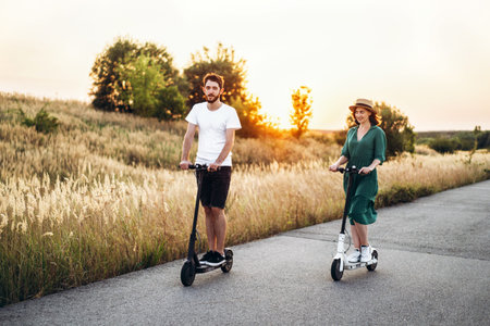 Happy couple walking on scooters. A young woman in dress and hat on sunset background. In the...