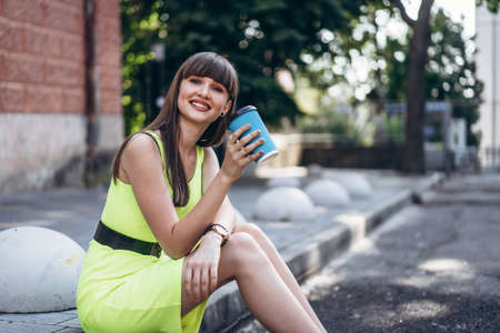 Pretty long hair brunette girl in green light dress with blue paper cup of beverage sitting outdoor on the street in the cityの写真素材