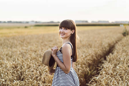 Pretty brunette girl in light dress wearing hat and walking outdoor in wheat field at sunset timeの写真素材