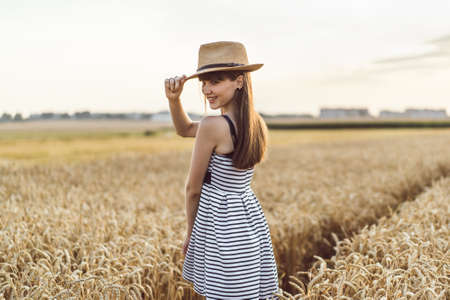 Pretty brunette girl in light dress wearing hat and walking outdoor in wheat field at sunset timeの写真素材