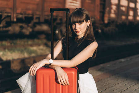 Female brunette traveler with red suitcase in white skirt waiting for a train on raiway stationの写真素材