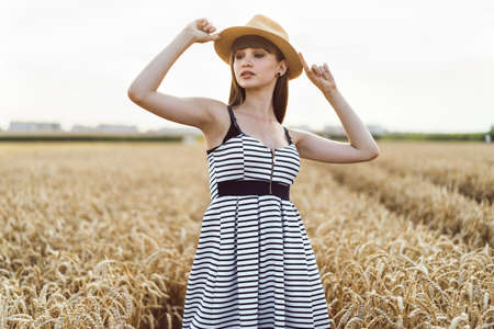 Pretty brunette girl in light dress wearing hat and walking outdoor in wheat field at sunset timeの写真素材