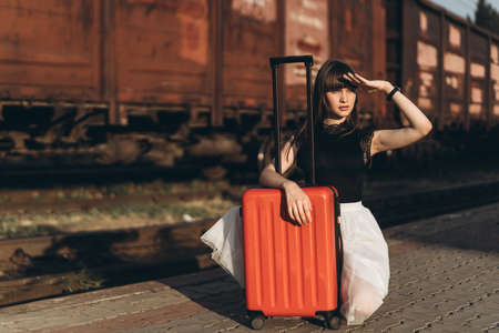 Female brunette traveler with red suitcase in white skirt waiting for a train on raiway stationの写真素材