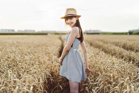 Pretty brunette girl in light dress wearing hat and walking outdoor in wheat field at sunset timeの写真素材