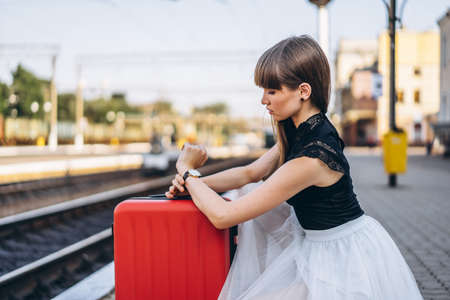 Female brunette traveler with red suitcase in white skirt waiting for a train on raiway station and checking time on her wristwatchの写真素材
