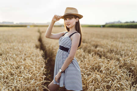 Pretty brunette girl in light dress wearing hat and walking outdoor in wheat field at sunset timeの写真素材