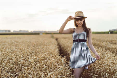 Pretty brunette girl in light dress wearing hat and walking outdoor in wheat field at sunset timeの写真素材