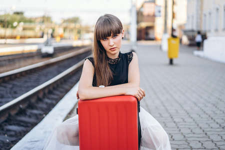 Female brunette traveler with red suitcase in white skirt waiting for a train on raiway stationの写真素材