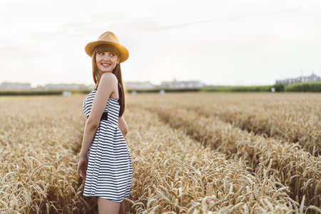 Pretty brunette girl in light dress wearing hat and walking outdoor in wheat field at sunset timeの写真素材