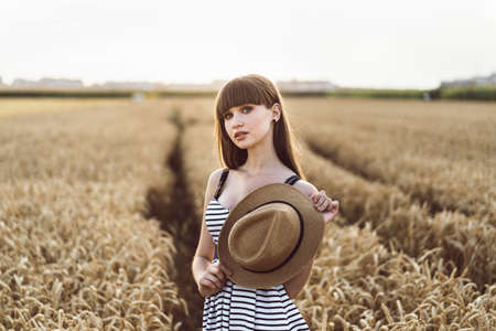 Pretty brunette girl in light dress wearing hat and walking outdoor in wheat field at sunset timeの写真素材