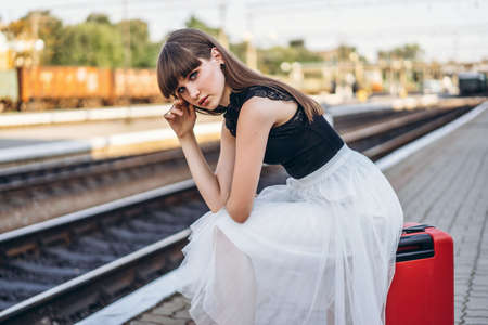 Female brunette traveler with red suitcase in white skirt waiting for a train on raiway stationの写真素材