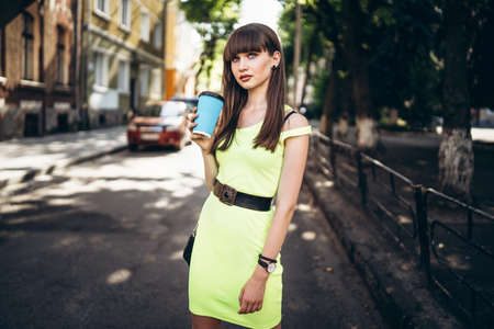 Pretty long hair brunette girl in green light dress with blue paper cup of beverage walking outdoor on the street in the cityの写真素材