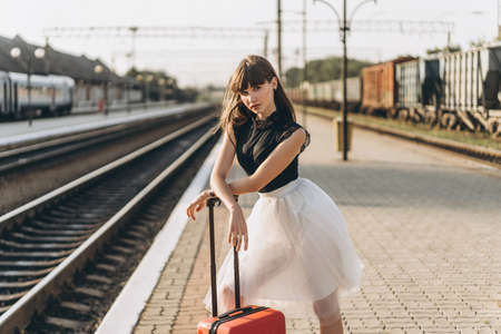 Female brunette traveler with red suitcase in white skirt waiting for a train and walking on raiway stationの写真素材