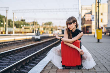 Female brunette traveler with red suitcase in white skirt waiting for a train on raiway station and checking time on her wristwatchの写真素材