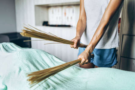 Female masseur in gray t-shirt and blue shorts doing massage with bamboo massage broom, client which is covered with large towel that lies on the massage couch, close-up of hand, concept of healthの写真素材