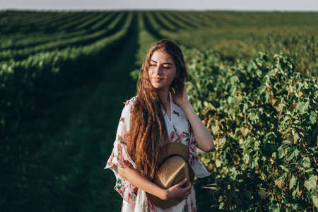 beautiful woman with long curly hair and freckles face on currant field background. Girl in a light dress walks in the summer sunny day.の写真素材