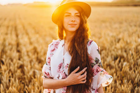 Portrait of a beautiful young woman with curly hair and freckles face. Woman in dress and hat posing in wheat field at sunset and looking at camera.の写真素材