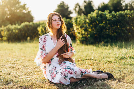 Full length portrait of beautiful woman with long curly hair on currant field background. Girl in a light dress sits on the grass in sunny day.の写真素材