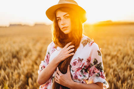 Portrait of a beautiful young woman with curly hair and freckles face. Woman in dress and hat posing in wheat field at sunset and looking at camera.の写真素材