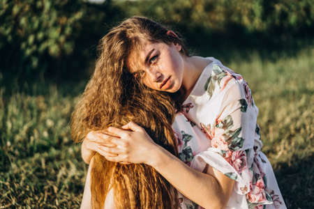 Young girl with long curly hair, brunette, face in freckles, with makeup and green eyes, in light white dress posing for camera, background blurred, natureの写真素材