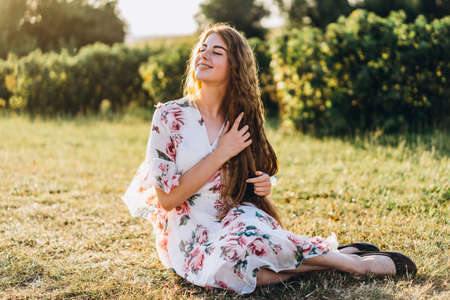 Full length portrait of beautiful woman with long curly hair on currant field background. Girl in a light dress sits on the grass in sunny day.の写真素材