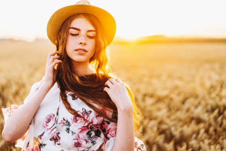 Portrait of a beautiful young woman with curly hair and freckles face. Woman in dress and hat posing in wheat field at sunset and looking at camera.の写真素材