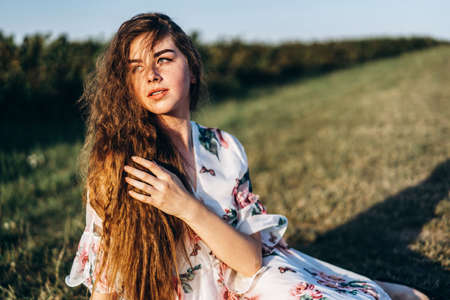 Young girl with long curly hair, brunette, face in freckles, with makeup and green eyes, in light white dress posing for camera, background blurred, natureの写真素材