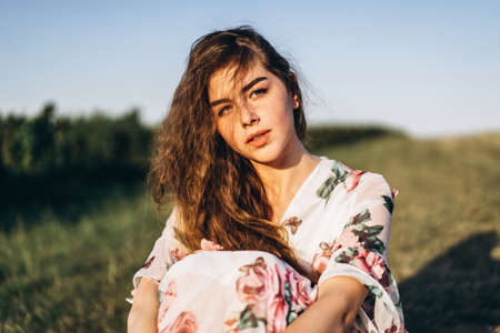 Young girl with long curly hair, brunette, face in freckles, with makeup and green eyes, in light white dress posing for camera, background blurred, natureの写真素材