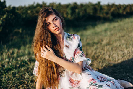 Young girl with long curly hair, brunette, face in freckles, with makeup and green eyes, in light white dress posing for camera, background blurred, natureの写真素材