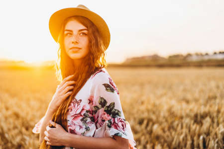 Portrait of a beautiful young woman with curly hair and freckles face. Woman in dress and hat posing in wheat field at sunset and looking at camera.の写真素材